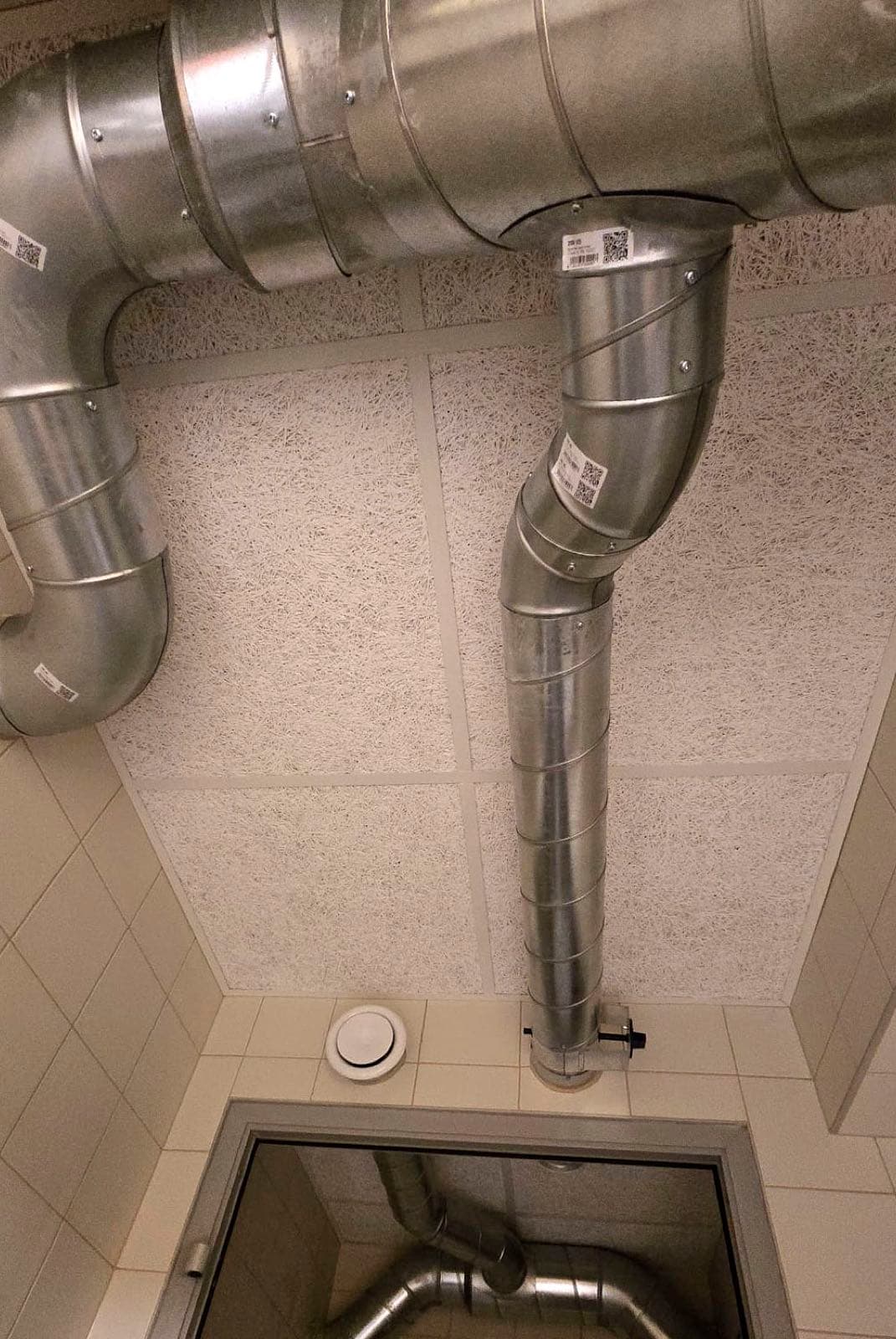 Baltas koka skaidu plāksnes nelielā telpā ar redzamiem ventilācijas cauruļvadiem